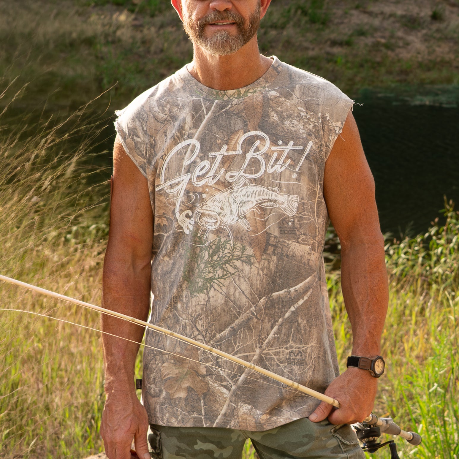 Man holding a fishing rod in a natural setting wearing a sleeveless camouflage shirt with "GET BIT" text.