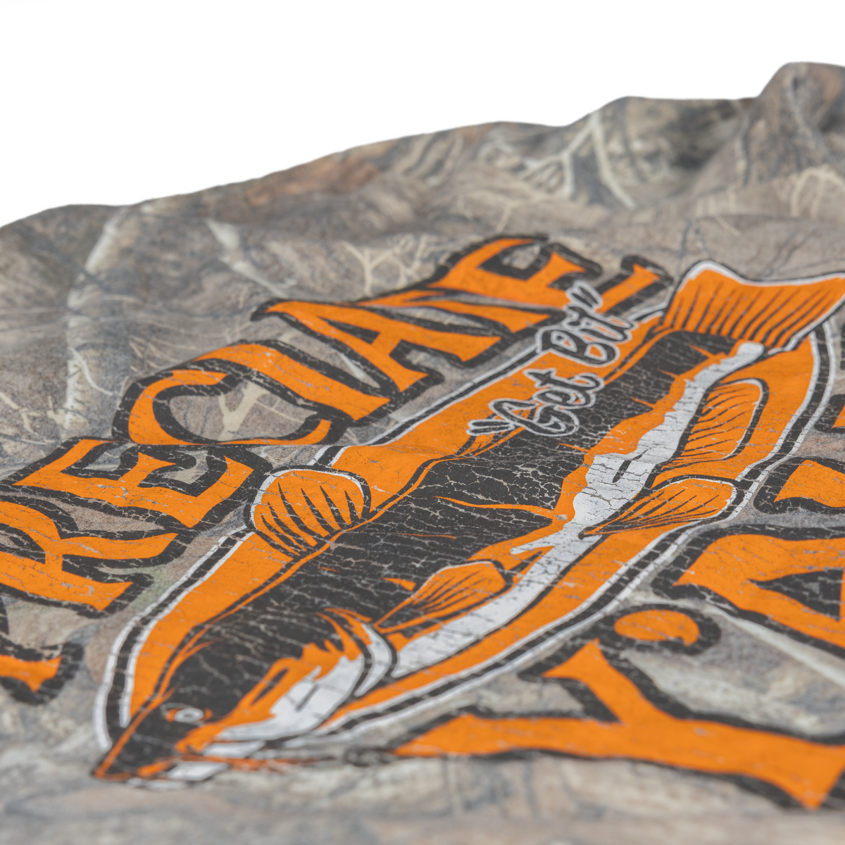 A close-up of a camouflage-patterned fabric surface with a large orange, black, and white fish graphic and stylized text printed across it.