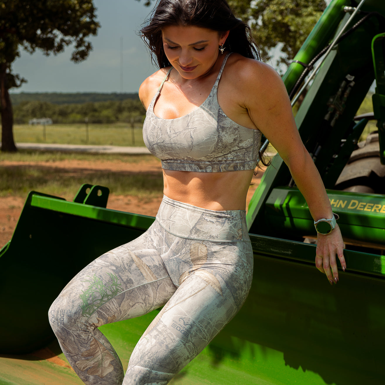 Woman in camouflage Leggings and sports bra standing on a green vehicle with trees in the background