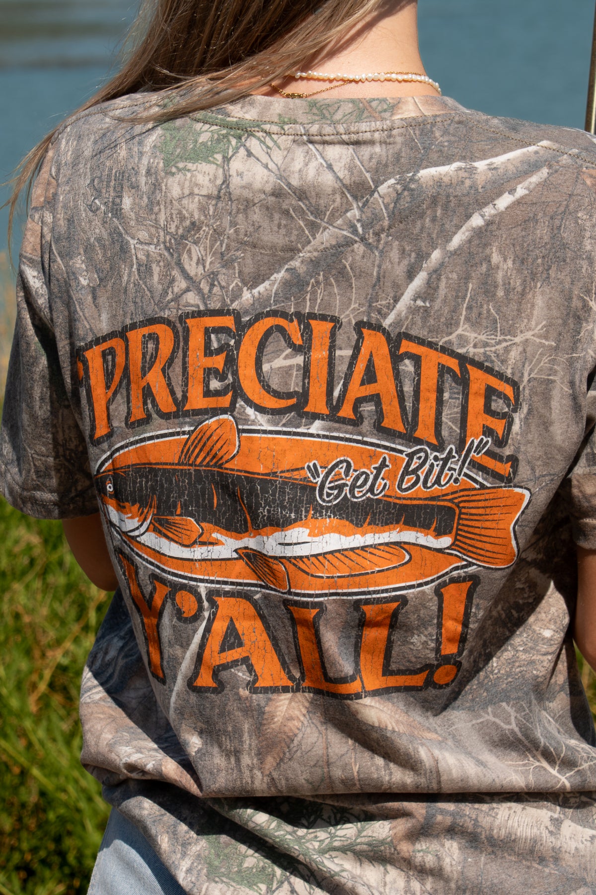 A woman stands outdoors by a lake wearing a camouflage graphic T-shirt with orange text reading "'PRECIATE Y'ALL" across the back along with fish graphic