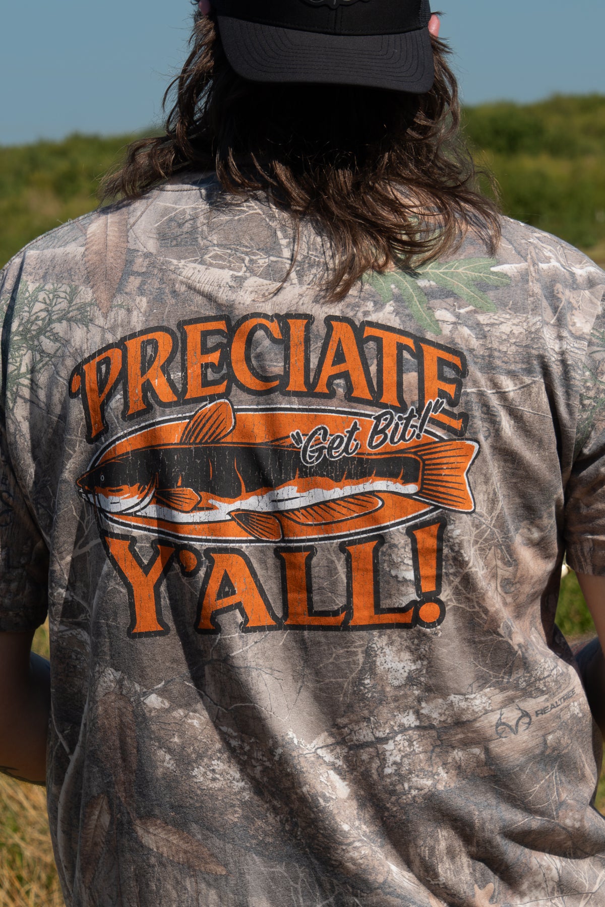 Man standing outside in camo tee with fish graphic on back with text "'PRECIATE Y'ALL! Get Bit!"