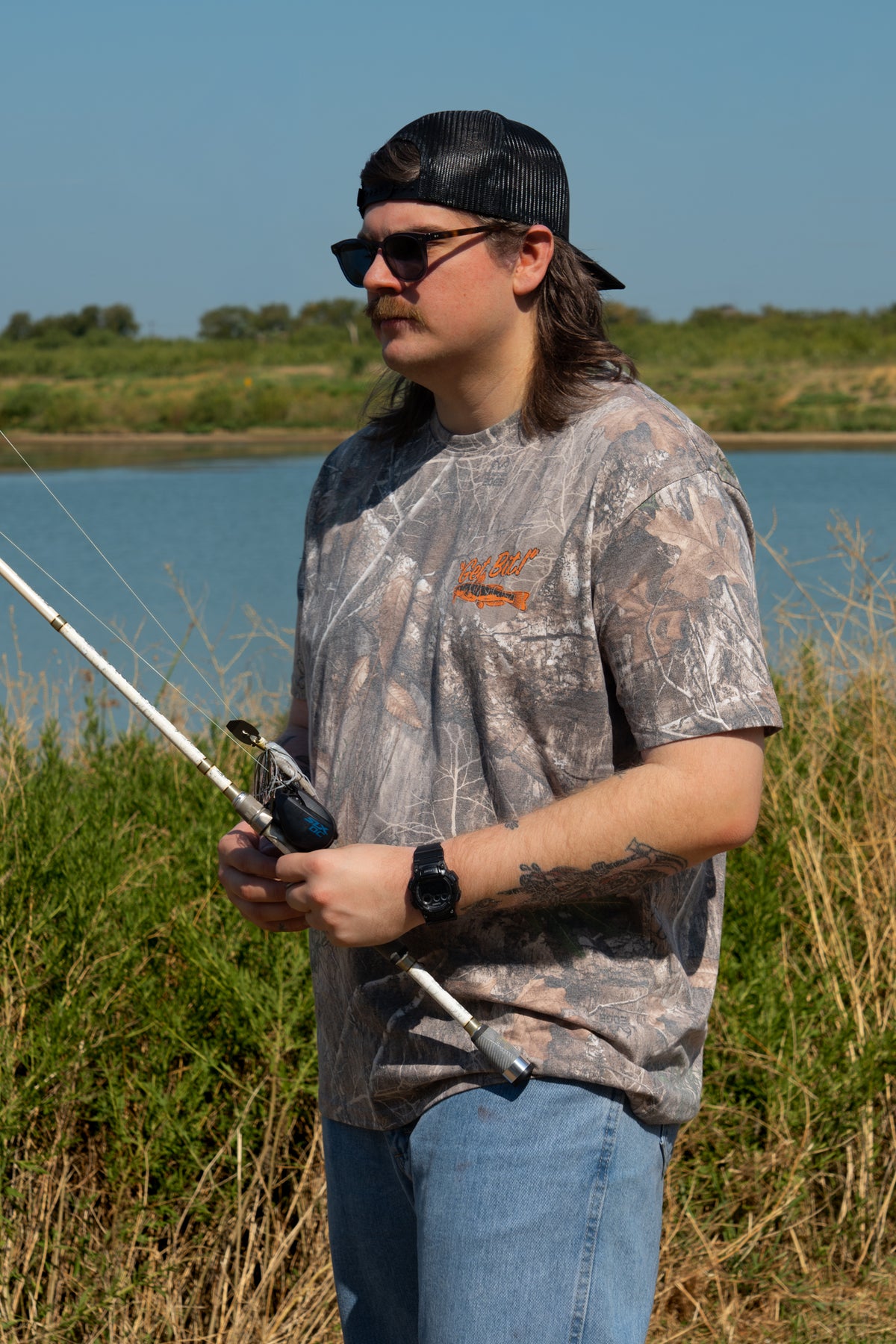 Man holding fishing pole wearing camo tee with fish and "Get Bit!" text on left chest