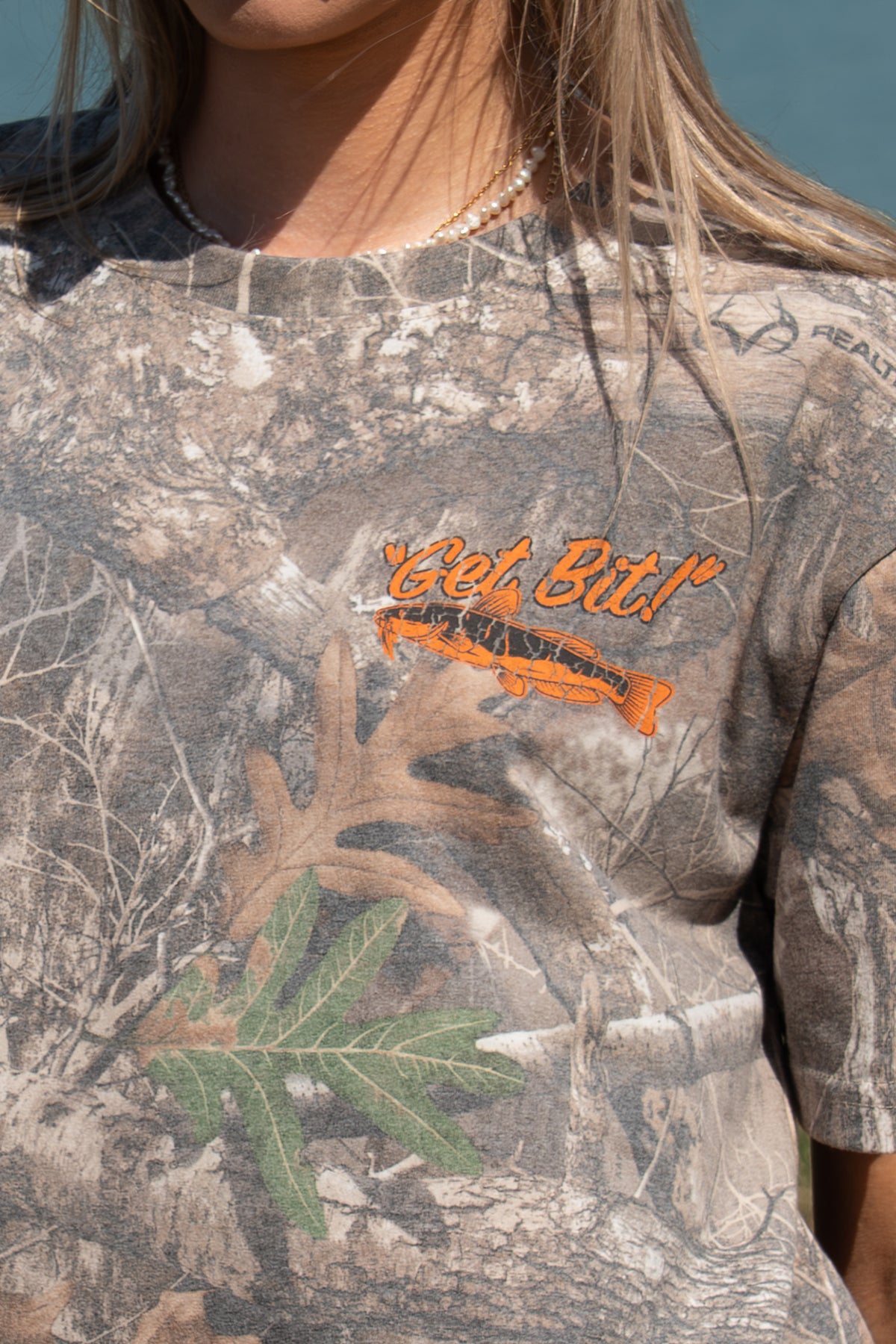 closeup of left chest of camo tee showing fish graphic with "Get Bit!" text
