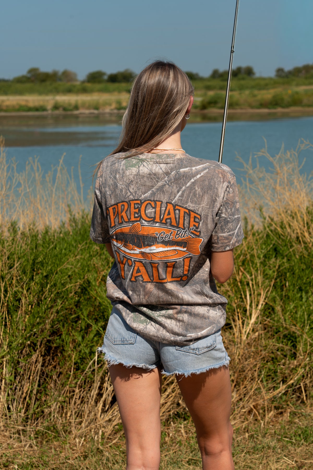 A woman stands outdoors by a lake wearing a camouflage graphic T-shirt with orange text reading "'PRECIATE Y'ALL" across the back along with fish graphic