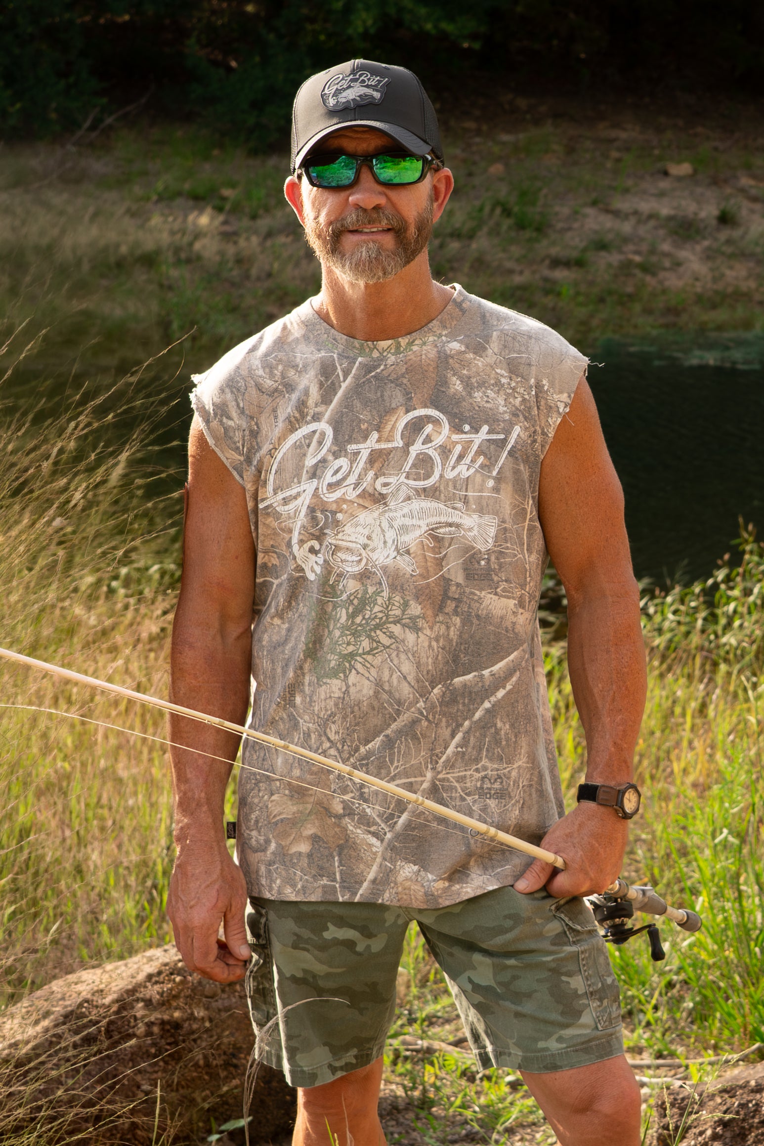 Man holding a fishing rod in a natural setting wearing a sleeveless camouflage shirt with "GET BIT" text.