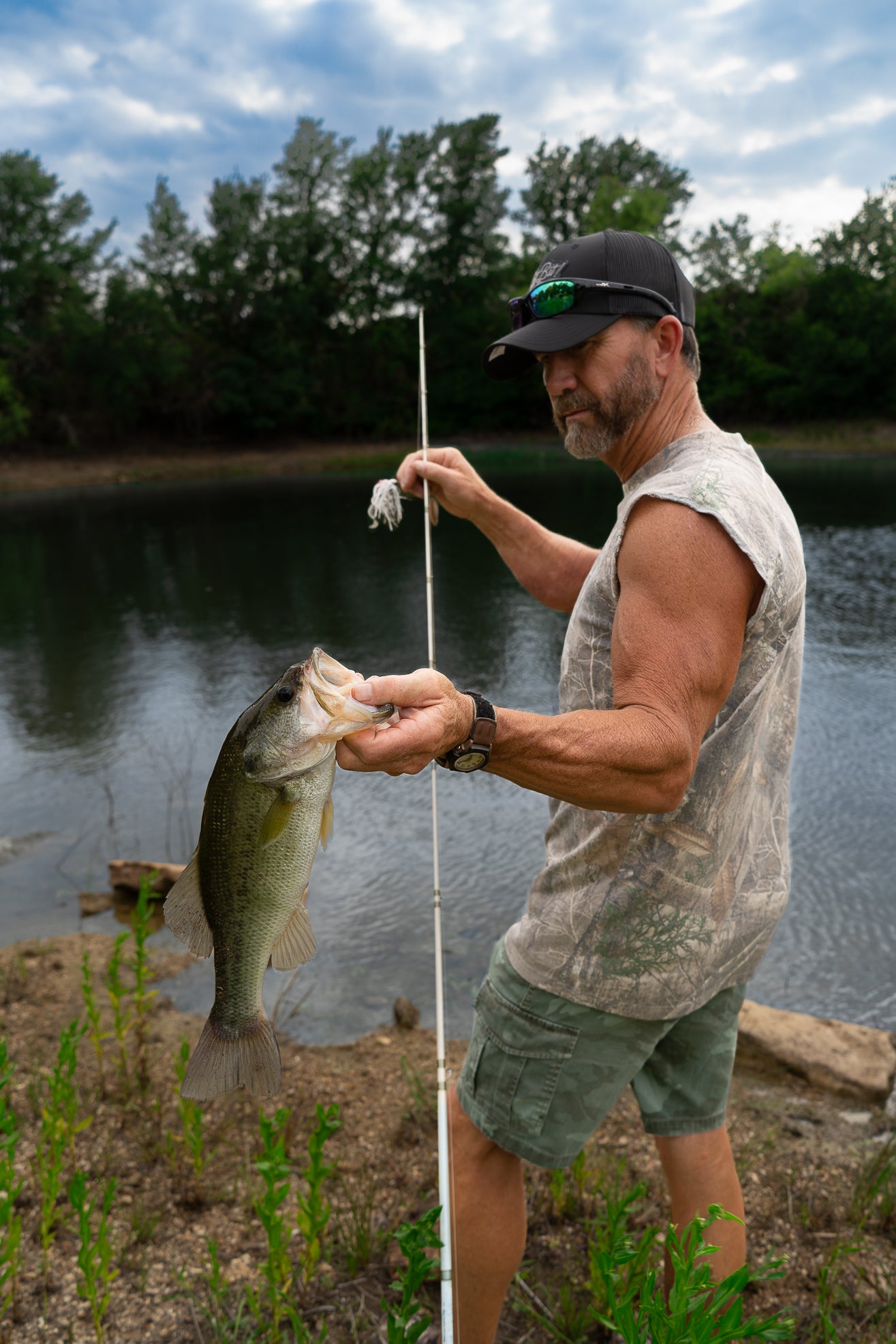 Man holding a fish caught on a fishing rod by a lake wearing camo shirt