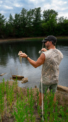 Man holding a fish by a lake with trees in the background wearing camo shirt