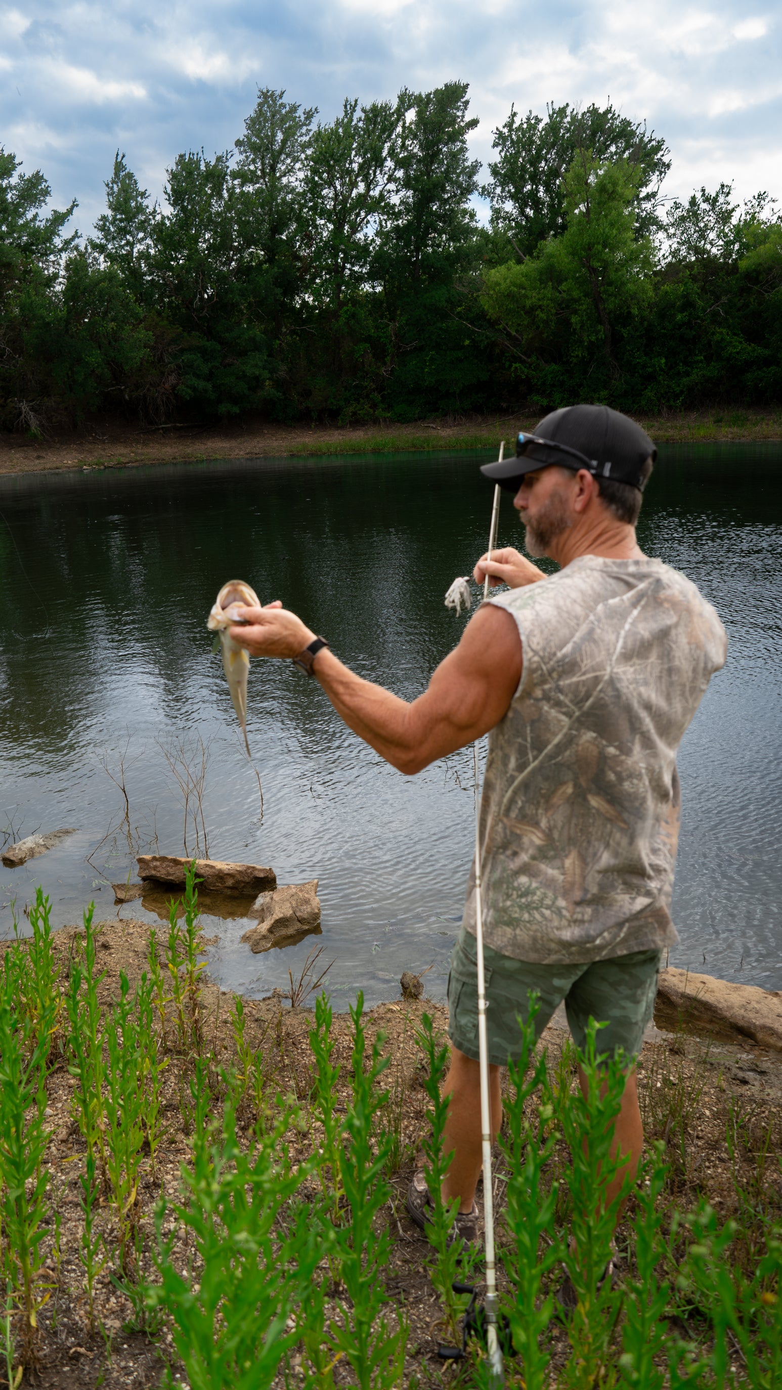 Man holding a fish by a lake with trees in the background wearing camo shirt