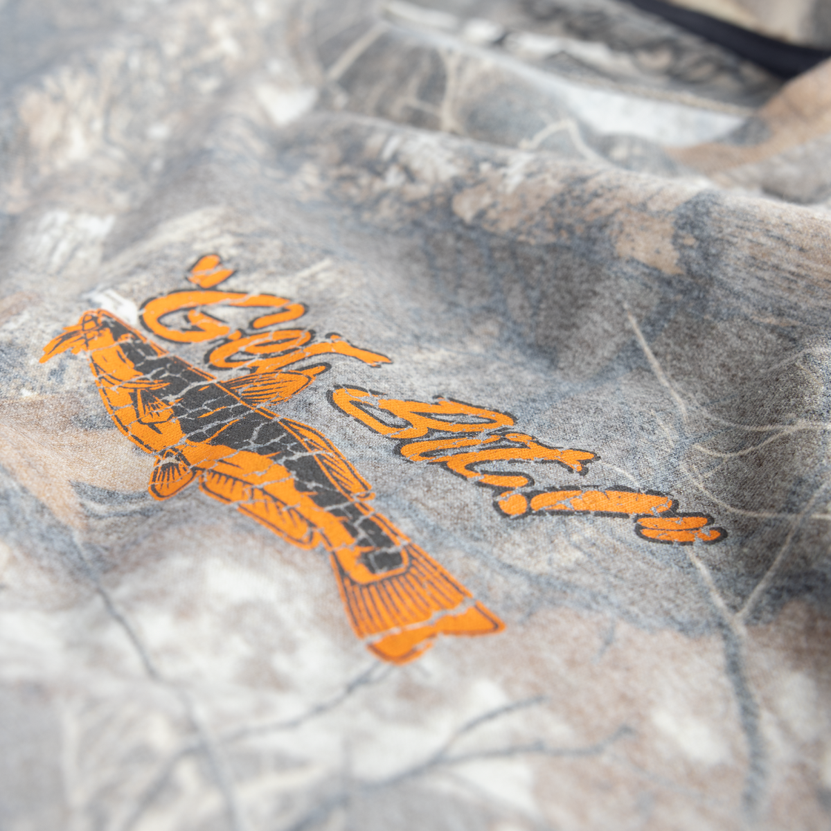 Close-up of a camouflage-patterned fabric with an orange and black fish graphic and the words "Get Bit!" in orange script.