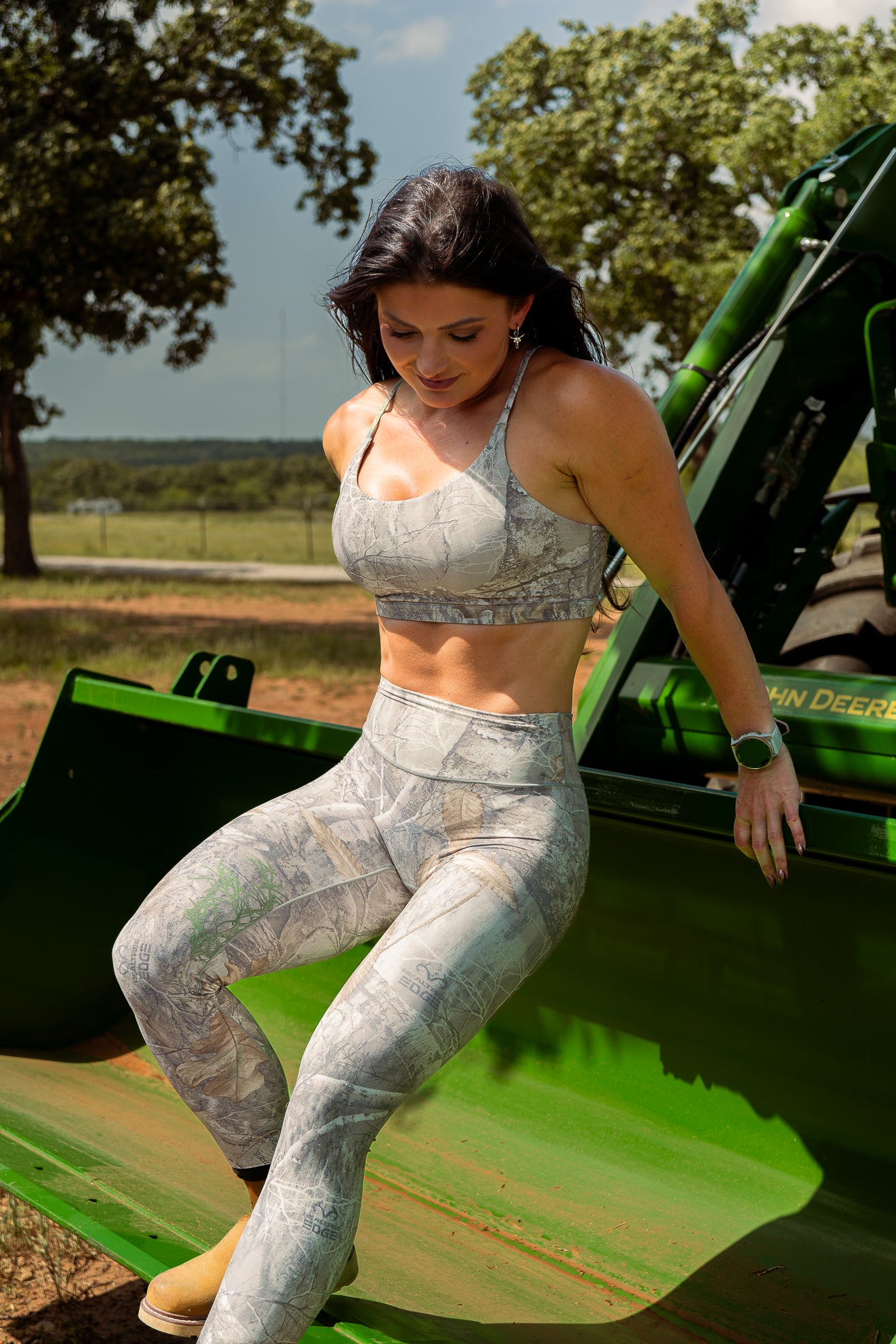 Woman in camouflage Leggings and sports bra standing on a green vehicle with trees in the background