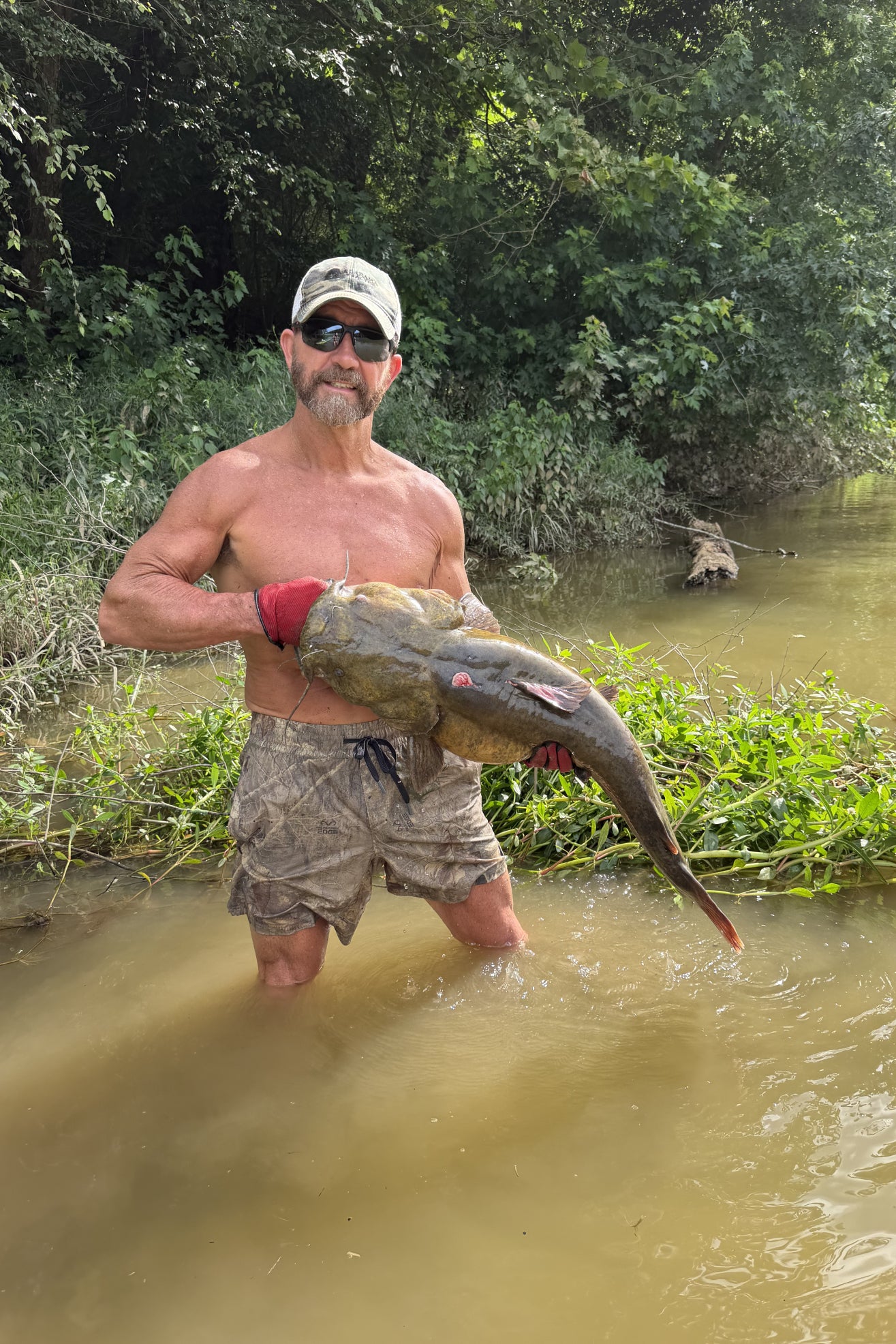 Man holding a large fish in a river wearing camo swim trunks