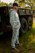 Man in camouflage gear standing next to a large truck in a grassy area.