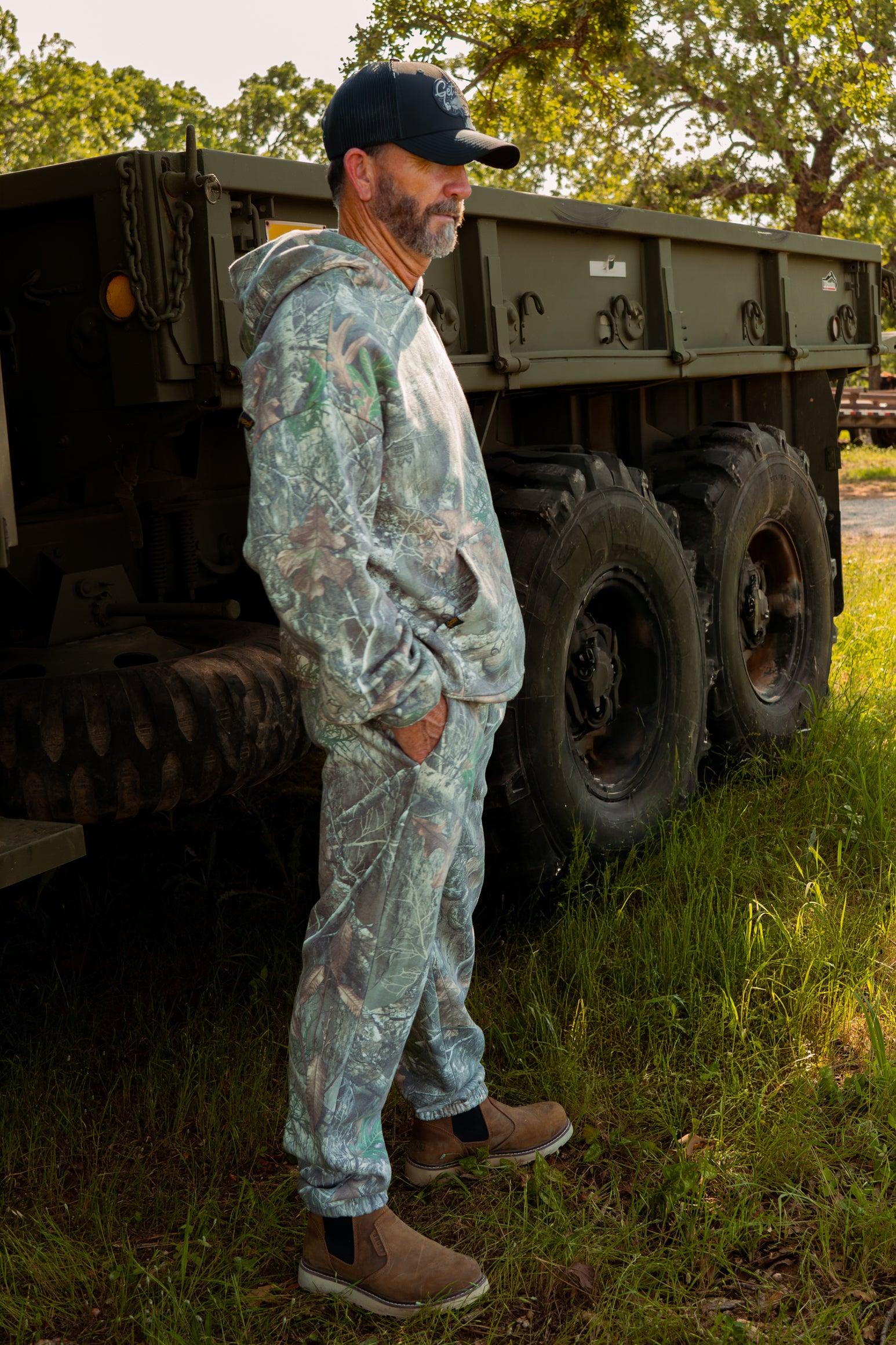 Man in camouflage gear standing next to a large truck in a grassy area.