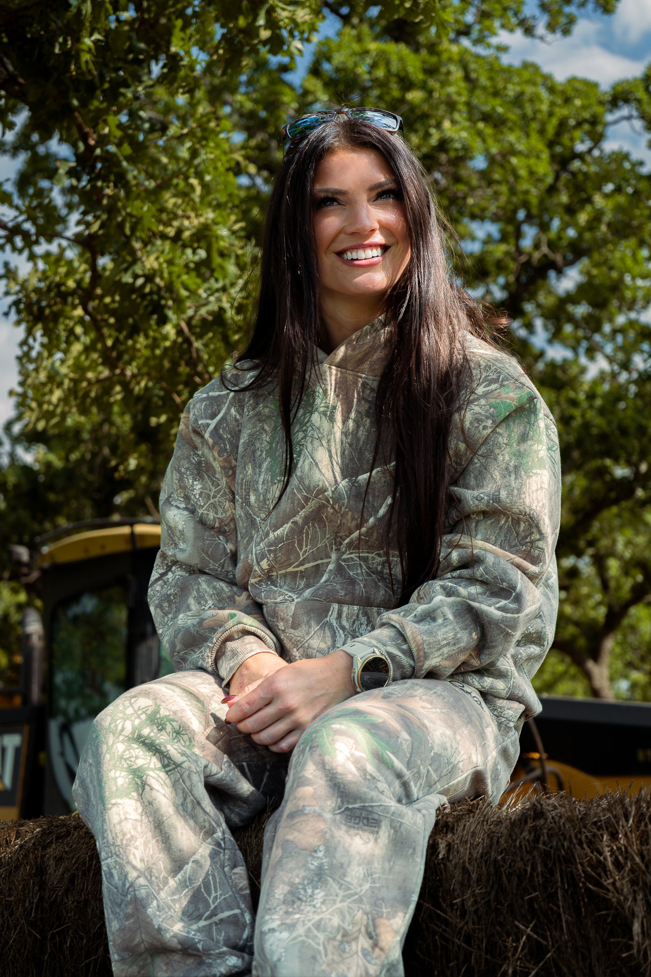 Woman in camouflage outfit sitting on a hay bale with trees in the background