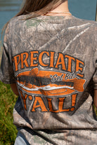 A woman stands outdoors by a lake wearing a camouflage graphic T-shirt with orange text reading "'PRECIATE Y'ALL" across the back along with fish graphic