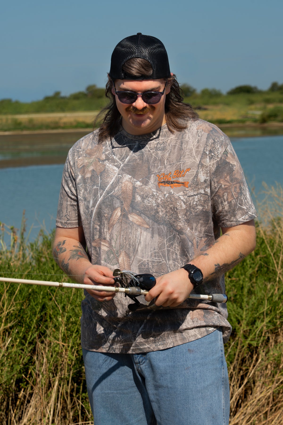 Man holding fishing pole wearing camo tee with fish on left chest with text "GET BIT!"