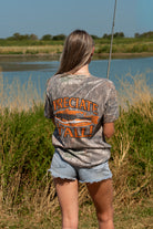 A woman stands outdoors by a lake wearing a camouflage graphic T-shirt with orange text reading "'PRECIATE Y'ALL" across the back along with fish graphic